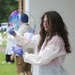 A person wearing a white disposable lab coat, blue nitrile gloves, safety goggles, and a wristwatch is manipulating a large, colorful soap bubble outdoors. The bubble displays vivid iridescent colors including blue, purple, and pink. In the background, other individuals in similar white protective gear are visible, suggesting a group science demonstration or educational activity.