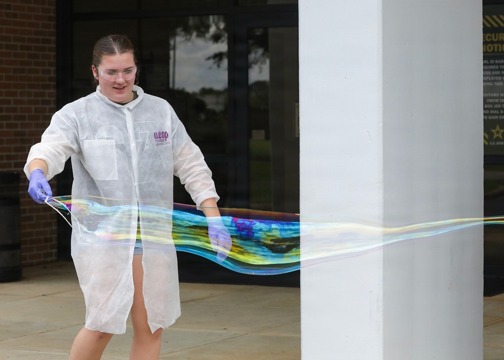 A young woman wearing a white disposable lab coat, safety goggles, and purple latex gloves is creating a large, iridescent soap bubble film outdoors in front of a building. She is stretching the bubble membrane between two wands, producing a wide, colorful, translucent sheet with rainbow-like interference colors. The setting appears to be a science demonstration or educational outreach event.