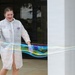 A young woman wearing a white disposable lab coat, safety goggles, and purple latex gloves is creating a large, iridescent soap bubble film outdoors in front of a building. She is stretching the bubble membrane between two wands, producing a wide, colorful, translucent sheet with rainbow-like interference colors. The setting appears to be a science demonstration or educational outreach event.