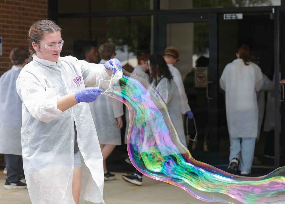 A young person wearing a white lab coat, safety goggles, and purple nitrile gloves is manipulating a large, iridescent soap bubble or thin film outdoors in front of a building. The bubble displays vivid rainbow colors including green, purple, blue, and orange. In the background, a group of similarly dressed individuals in white lab coats are entering or standing near glass doors of a brick building.