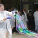 A young person wearing a white lab coat, safety goggles, and purple nitrile gloves is manipulating a large, iridescent soap bubble or thin film outdoors in front of a building. The bubble displays vivid rainbow colors including green, purple, blue, and orange. In the background, a group of similarly dressed individuals in white lab coats are entering or standing near glass doors of a brick building.