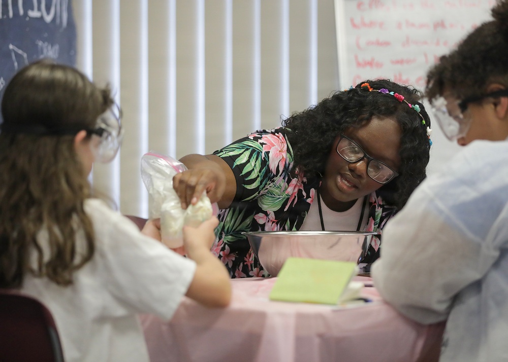 A group of students, likely in a science class or educational workshop, are engaged in a hands-on experiment at a table covered with a pink tablecloth. A young woman wearing glasses and a floral shirt appears to be instructing or assisting two other students with a bag containing a white substance over a metal bowl. Safety goggles are visible on at least two participants, indicating a structured science activity.