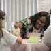 A group of students, likely in a science class or educational workshop, are engaged in a hands-on experiment at a table covered with a pink tablecloth. A young woman wearing glasses and a floral shirt appears to be instructing or assisting two other students with a bag containing a white substance over a metal bowl. Safety goggles are visible on at least two participants, indicating a structured science activity.
