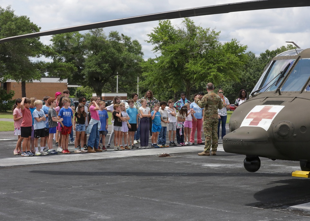 A uniformed soldier in camouflage (OCP) uniform is briefing a group of children and adult chaperones beside a military medical evacuation (MEDEVAC) helicopter, identifiable by the red cross markings on its olive-drab fuselage. The setting appears to be a school or community facility parking lot during an educational outreach or career day event. The helicopter's main rotor blade is visible overhead, framing the group.