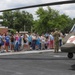 A uniformed soldier in camouflage (OCP) uniform is briefing a group of children and adult chaperones beside a military medical evacuation (MEDEVAC) helicopter, identifiable by the red cross markings on its olive-drab fuselage. The setting appears to be a school or community facility parking lot during an educational outreach or career day event. The helicopter's main rotor blade is visible overhead, framing the group.