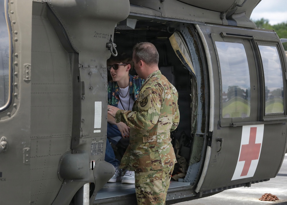 A military service member in OCP (Operational Camouflage Pattern) uniform is speaking with a civilian visitor seated inside a military medical helicopter, identifiable by the red cross markings on its fuselage. The interaction appears to be an informational tour or demonstration, likely at a public event or open house. The helicopter is a UH-60 Black Hawk configured for medevac/CASEVAC operations.