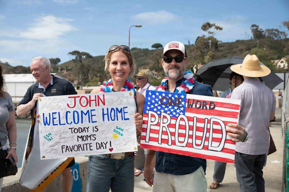 Two adults are holding handmade welcome home signs at what appears to be a military homecoming event, likely at a naval base in San Diego. The woman holds a sign reading 'JOHN WELCOME HOME – TODAY IS MOM'S FAVORITE DAY!' with 'SSN 761' and 'SAN DIEGO' noted, suggesting a submarine crew member is returning. The man holds an American flag-themed sign reading 'ONE WORD: PROUD', and both are wearing patriotic leis.