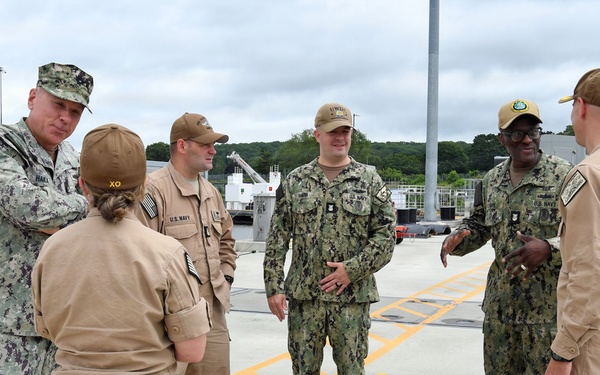 A group of six U.S. Navy personnel are gathered in a circle on what appears to be a naval facility dock or pier, engaged in conversation. The personnel are wearing a mix of Navy Working Uniform (NWU Type III camouflage) and tan/khaki Navy coveralls or utilities. The setting appears to be an active waterfront installation with naval vessels visible in the background.