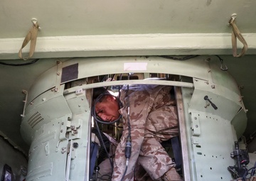 A soldier in desert camouflage uniform is working inside a military armored vehicle, appearing to perform maintenance or inspection tasks. The interior shows a cramped space with various cables, electronic equipment, and military hardware. The vehicle interior is painted in a light olive/green color typical of military vehicles.