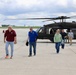 A group of civilians and at least one military personnel are walking away from a black military helicopter, likely a UH-60 Black Hawk, on a concrete tarmac. The setting appears to be a military or joint-use airfield, with an air traffic control tower visible in the background. The group appears to have just disembarked from the helicopter.