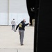 The image shows an outdoor tarmac or airfield apron area near a large hangar structure. A man in the foreground wearing a black hoodie, gray cargo pants, sunglasses, and aviation headset is giving a thumbs-up gesture toward what appears to be an aircraft (partially visible on the right). Two other individuals are visible in the background near the hangar.