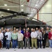 A large group of approximately 35-40 individuals poses for a group photograph inside a military aviation hangar in front of a dark-colored UH-60 Black Hawk helicopter. The group includes a mix of military personnel in Army Combat Uniforms (ACUs) and civilians wearing business casual attire, many with visible lanyards and badges. The setting appears to be an official visit or tour of a military aviation facility.