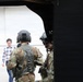 The image is taken from inside a military aircraft, looking outward through an open door or hatch. Two military personnel in camouflage uniforms and flight helmets are visible in the foreground, while a civilian man in a plaid shirt stands in the background on what appears to be a tarmac or airfield apron. The scene suggests a briefing, demonstration, or official visit at a military aviation facility.