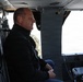 A middle-aged man wearing a black jacket and jeans is seated inside what appears to be a military helicopter, likely as a civilian passenger or observer. He is wearing an orange ear plug and appears to be looking forward calmly. In the background, a person in military camouflage uniform is partially visible near an open door.