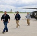 Three civilians wearing visitor badges walk across a military airfield tarmac, appearing to have just disembarked from or are walking near military helicopters. Two dark military helicopters, consistent with UH-60 Black Hawks, are visible on the tarmac. At least one uniformed military personnel member is visible near the right helicopter, suggesting this may be an official visit or demonstration event.