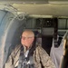 An older bald man wearing camouflage military-style clothing and glasses is seated inside what appears to be a military helicopter, secured with a harness/seatbelt system. He is looking out the window to his left, suggesting the aircraft is in flight. The interior shows typical military rotary-wing aircraft features including padded walls, straps, and structural components.