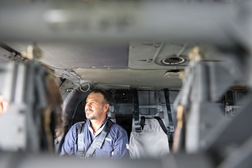 A middle-aged man wearing a dark blue work uniform with a chest harness is seated inside what appears to be a military or utility helicopter cabin. The photo is taken through an opening or doorway, creating a framed, depth-of-field effect with blurred foreground elements. The man appears to be looking to his left, possibly observing something outside the aircraft.