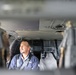 A middle-aged man wearing a dark blue work uniform with a chest harness is seated inside what appears to be a military or utility helicopter cabin. The photo is taken through an opening or doorway, creating a framed, depth-of-field effect with blurred foreground elements. The man appears to be looking to his left, possibly observing something outside the aircraft.