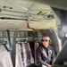 A man is seated inside what appears to be a military or utility helicopter cabin, wearing blue mirrored sunglasses and a communications headset. He is secured with a harness/seatbelt system and dressed in a dark sweatshirt. The interior shows typical rotary-wing aircraft features including fold-down troop seats, warning placards, and overhead equipment.