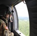 A U.S. Army soldier wearing a flight helmet and OCP (Operational Camouflage Pattern) uniform is seated inside a military helicopter, looking out an open side door at a forested landscape below. The soldier is equipped with a flight helmet featuring a visor, a communications headset, and tactical gear including a chest rig. The aerial perspective suggests the helicopter is at moderate altitude over a densely wooded area.