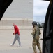 The image is taken from inside a military helicopter or aircraft, looking out through an open door onto a tarmac or airfield apron. A civilian man in a red jacket and gray pants is walking and smiling, while a military personnel in OCP (Operational Camouflage Pattern) uniform with a flight helmet and tactical vest stands nearby. A large industrial or hangar building is visible in the background.