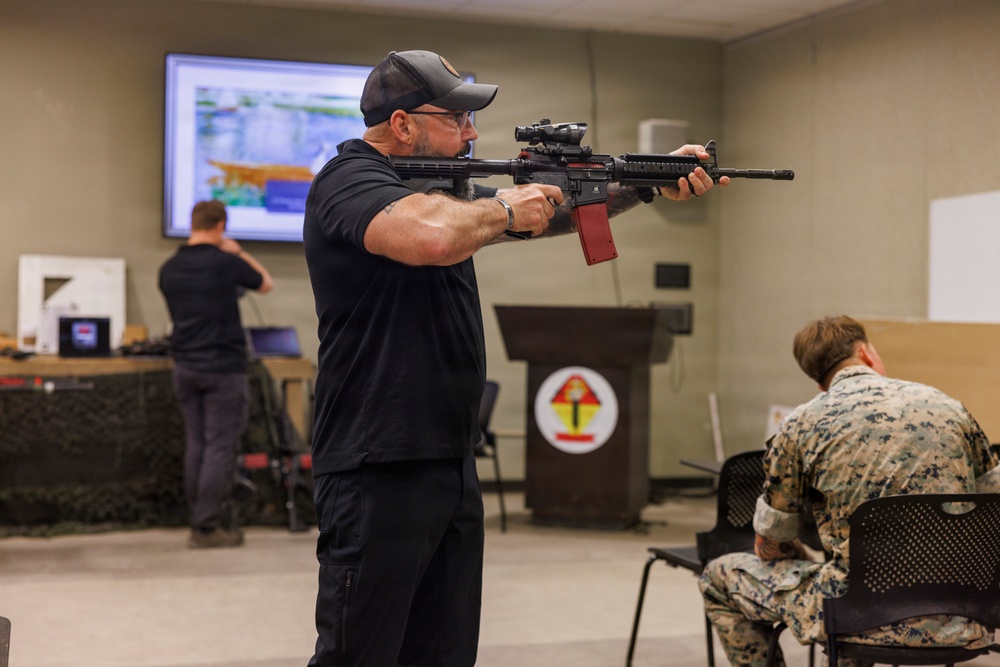 An indoor briefing or demonstration room shows a civilian instructor in black clothing and cap aiming an AR-style rifle with a red magazine and optical scope toward the left of the frame. A Marine in digital camouflage uniform sits to the right taking notes, while another person in black operates equipment at a table in the background. A podium bearing what appears to be a U.S. Marine Corps unit insignia is visible in the center background.