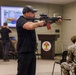 An indoor briefing or demonstration room shows a civilian instructor in black clothing and cap aiming an AR-style rifle with a red magazine and optical scope toward the left of the frame. A Marine in digital camouflage uniform sits to the right taking notes, while another person in black operates equipment at a table in the background. A podium bearing what appears to be a U.S. Marine Corps unit insignia is visible in the center background.