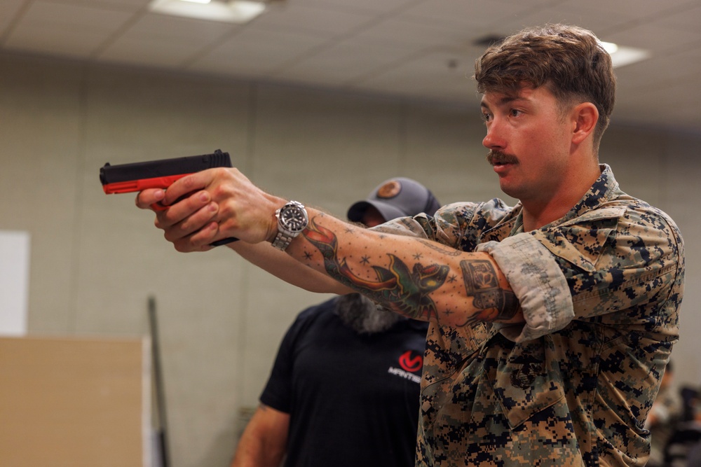 A young man in US Marine Corps MARPAT digital camouflage uniform is holding a training pistol (identified by its red/orange frame, indicating a non-lethal or inert training weapon) in a two-handed shooting stance. He has visible tattoos on his forearms and is wearing a wristwatch. The setting appears to be an indoor training facility or classroom environment.
