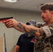 A young man in US Marine Corps MARPAT digital camouflage uniform is holding a training pistol (identified by its red/orange frame, indicating a non-lethal or inert training weapon) in a two-handed shooting stance. He has visible tattoos on his forearms and is wearing a wristwatch. The setting appears to be an indoor training facility or classroom environment.
