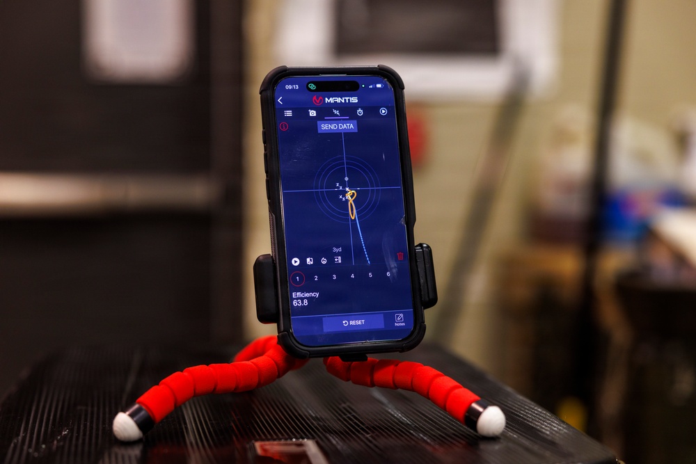 A smartphone displaying the MANTIS shooting training app is mounted on a flexible red tripod with white-tipped legs. The app screen shows a shot tracking interface with a radar-style display, trajectory path, and an efficiency score of 63.8. The phone is positioned on what appears to be a dark surface in an indoor environment.
