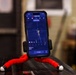 A smartphone displaying the MANTIS shooting training app is mounted on a flexible red tripod with white-tipped legs. The app screen shows a shot tracking interface with a radar-style display, trajectory path, and an efficiency score of 63.8. The phone is positioned on what appears to be a dark surface in an indoor environment.