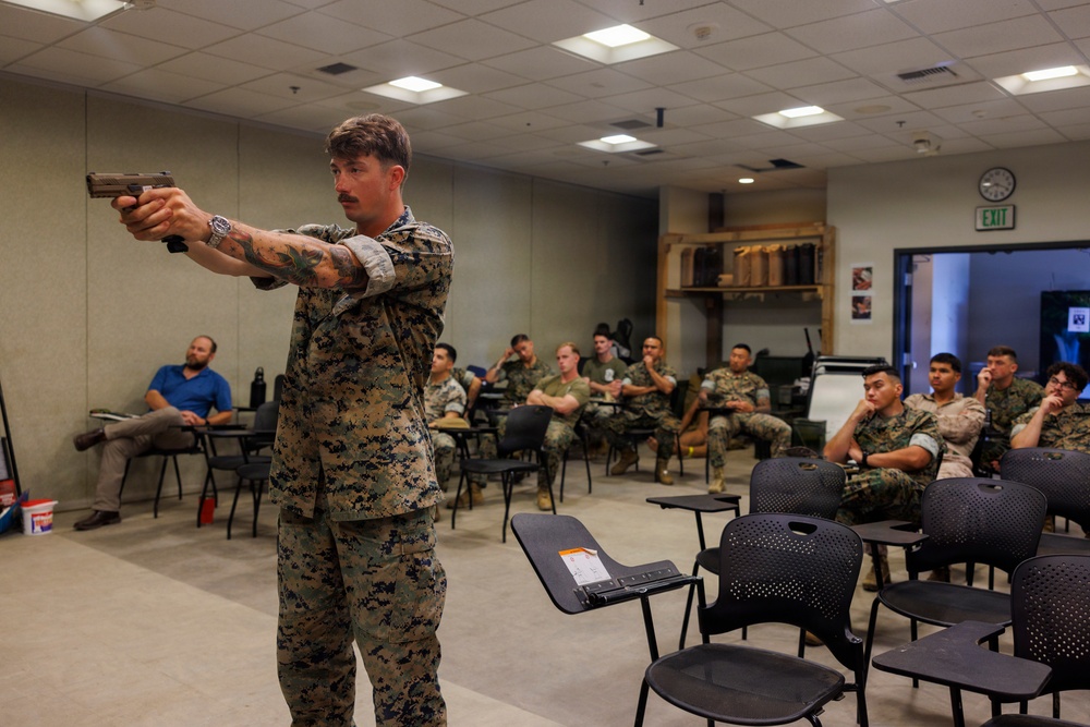 A military instructor in USMC digital camouflage uniform is demonstrating pistol handling technique to a classroom of seated Marines. The instructor, who has visible tattoos on his arms, is extending a handgun in a two-handed grip stance as part of what appears to be a weapons training or marksmanship instruction session. The audience of approximately 15-20 personnel in military uniforms are attentively observing the demonstration.