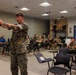 A military instructor in USMC digital camouflage uniform is demonstrating pistol handling technique to a classroom of seated Marines. The instructor, who has visible tattoos on his arms, is extending a handgun in a two-handed grip stance as part of what appears to be a weapons training or marksmanship instruction session. The audience of approximately 15-20 personnel in military uniforms are attentively observing the demonstration.