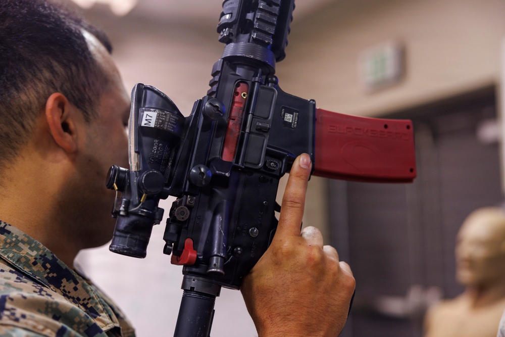 A person wearing U.S. Marine Corps MARPAT digital camouflage uniform is closely inspecting a training/simulation rifle equipped with a red magazine and an M7 laser training device attachment. The weapon appears to be a MILES (Multiple Integrated Laser Engagement System) or similar force-on-force training system, identifiable by the laser emitter and red training magazine. The setting appears to be an indoor training facility.
