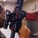 A person wearing U.S. Marine Corps MARPAT digital camouflage uniform is closely inspecting a training/simulation rifle equipped with a red magazine and an M7 laser training device attachment. The weapon appears to be a MILES (Multiple Integrated Laser Engagement System) or similar force-on-force training system, identifiable by the laser emitter and red training magazine. The setting appears to be an indoor training facility.