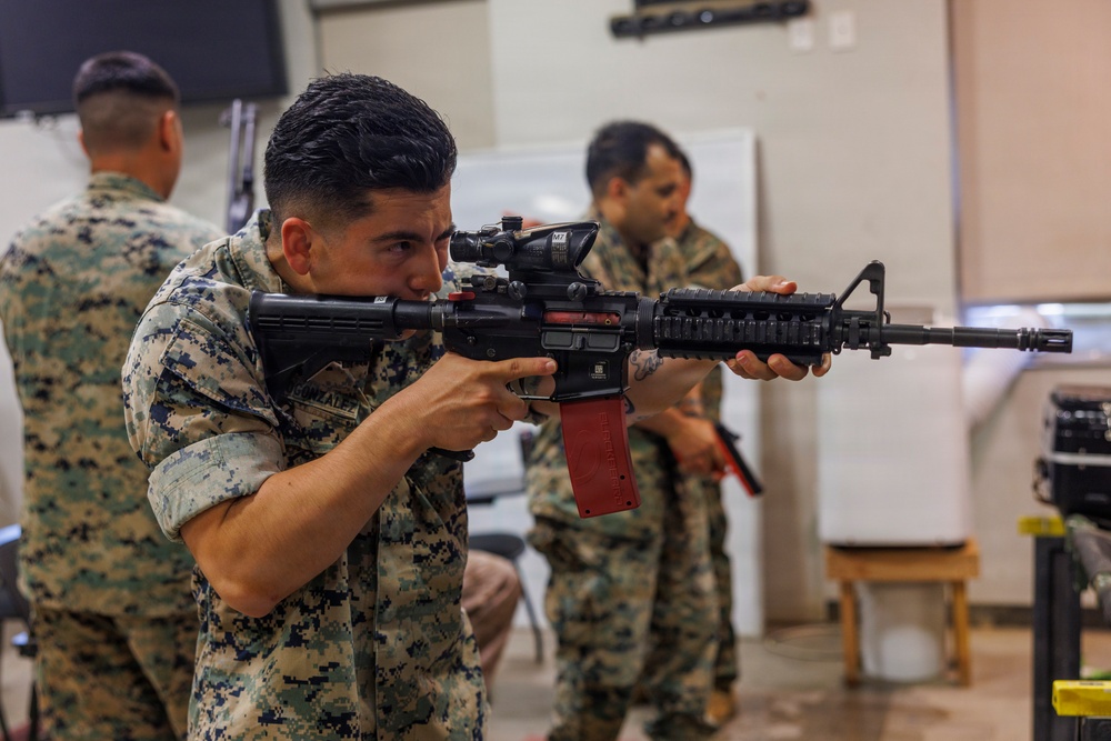 A U.S. Marine in MARPAT digital camouflage uniform is aiming a rifle with an optical scope and a red training magazine in an indoor facility. At least two other Marines in the same uniform are visible in the background. The setting appears to be a training or equipment demonstration environment.