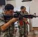 A U.S. Marine in MARPAT digital camouflage uniform is aiming a rifle with an optical scope and a red training magazine in an indoor facility. At least two other Marines in the same uniform are visible in the background. The setting appears to be a training or equipment demonstration environment.