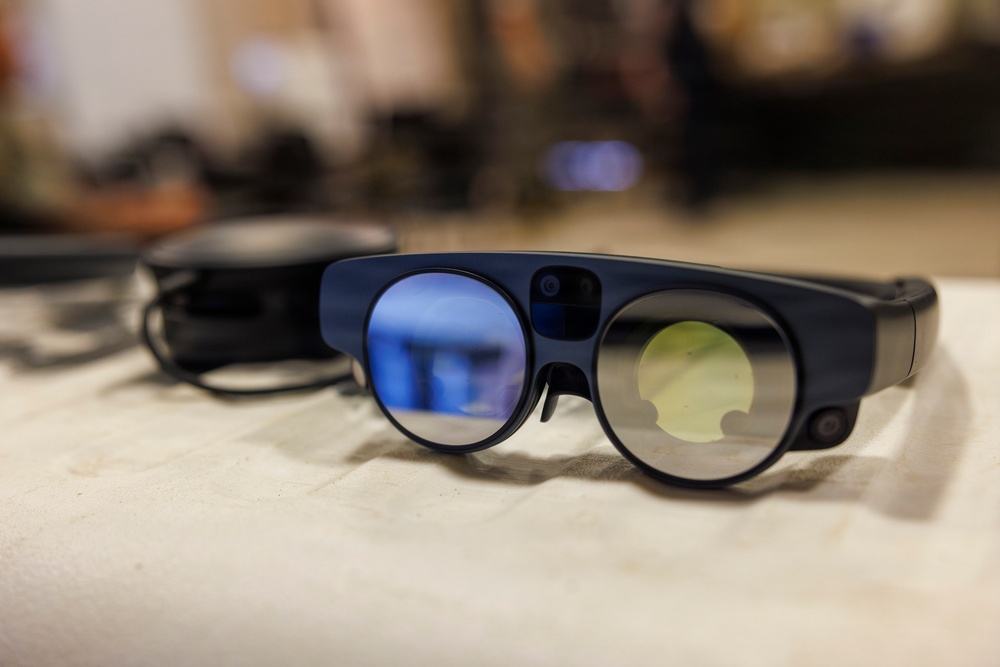 The image shows a pair of augmented reality (AR) smart glasses resting on a white fabric surface. The glasses feature two circular lenses with distinct blue and yellow-green tinted coatings, a thick dark blue frame, and what appears to be a small camera or sensor mounted on the bridge. A second, darker device is partially visible and out of focus in the background.