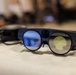 The image shows a pair of augmented reality (AR) smart glasses resting on a white fabric surface. The glasses feature two circular lenses with distinct blue and yellow-green tinted coatings, a thick dark blue frame, and what appears to be a small camera or sensor mounted on the bridge. A second, darker device is partially visible and out of focus in the background.