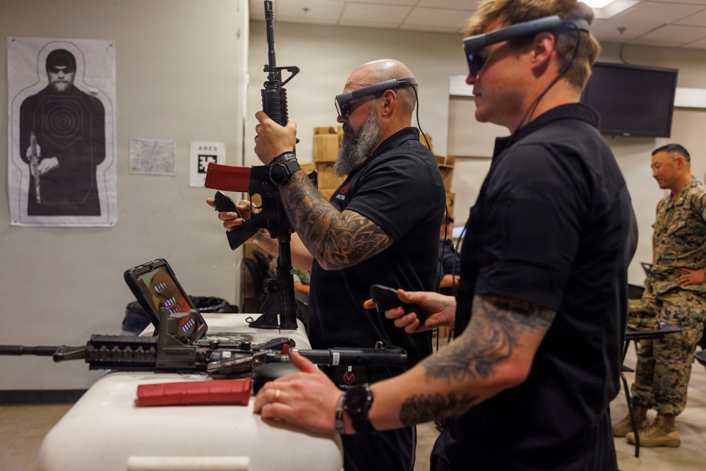 An indoor demonstration or training session featuring augmented reality (AR) headsets and simulated firearms. Two civilians in black shirts wearing AR/mixed reality glasses are handling what appears to be a rifle simulator, while military personnel in camouflage uniforms observe in the background. A shooting target poster is visible on the wall, suggesting this is a weapons training simulation technology demonstration.