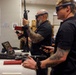 An indoor demonstration or training session featuring augmented reality (AR) headsets and simulated firearms. Two civilians in black shirts wearing AR/mixed reality glasses are handling what appears to be a rifle simulator, while military personnel in camouflage uniforms observe in the background. A shooting target poster is visible on the wall, suggesting this is a weapons training simulation technology demonstration.