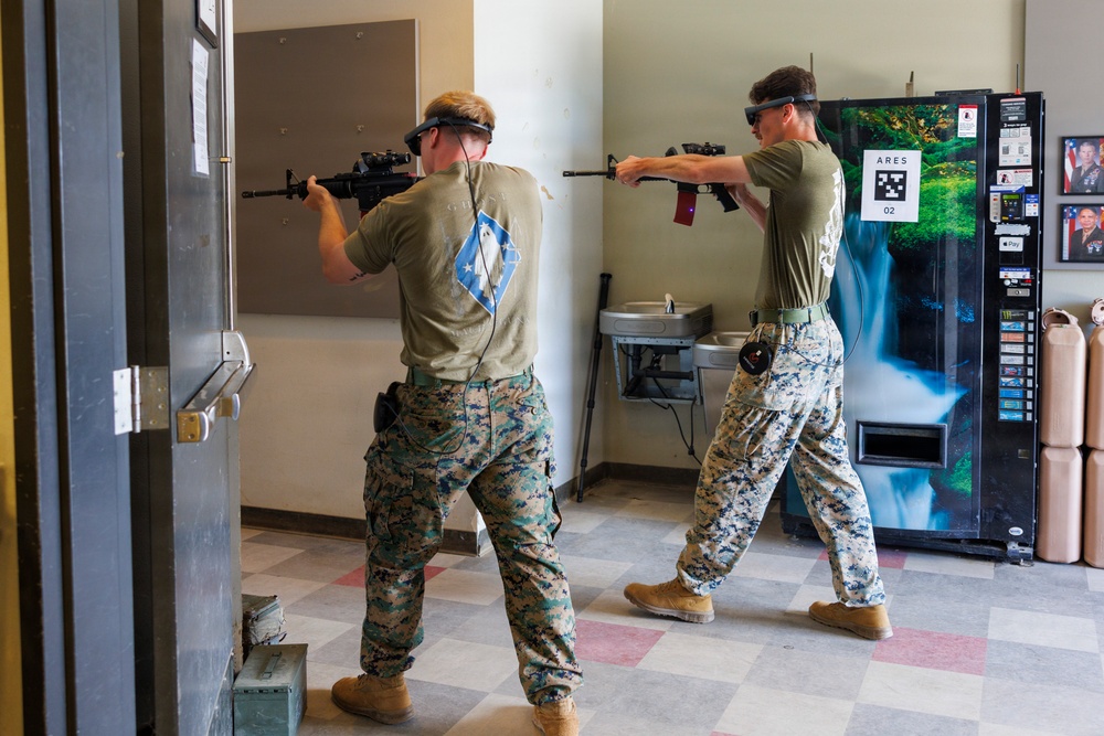 Two U.S. Marines in MARPAT camouflage uniforms are conducting an augmented reality (AR) training exercise indoors, wearing AR headsets and aiming rifles. The setting appears to be a military facility hallway or common area, with a vending machine and water fountain visible in the background. The system in use appears to be the ARES (Augmented Reality Enhanced Soldier) training platform, indicated by a QR-style marker on the vending machine.