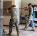 Two U.S. Marines in MARPAT camouflage uniforms are conducting an augmented reality (AR) training exercise indoors, wearing AR headsets and aiming rifles. The setting appears to be a military facility hallway or common area, with a vending machine and water fountain visible in the background. The system in use appears to be the ARES (Augmented Reality Enhanced Soldier) training platform, indicated by a QR-style marker on the vending machine.