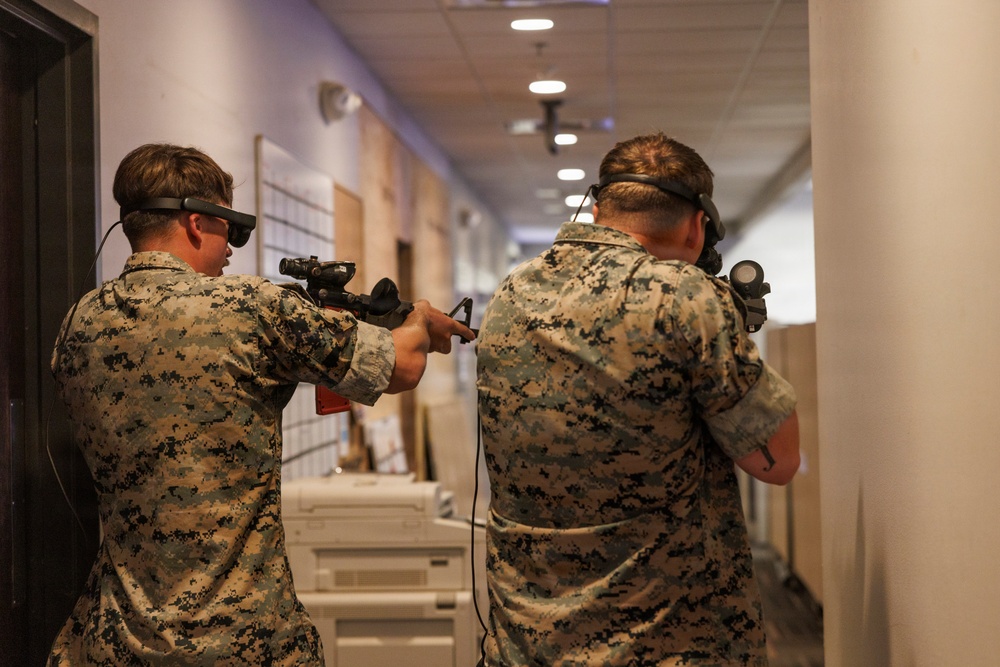 Two individuals wearing digital camouflage uniforms (consistent with USMC MARPAT pattern) are participating in what appears to be an augmented or mixed reality training exercise in an indoor corridor. Both are equipped with head-mounted display devices and are holding simulated weapon controllers with cables attached. The setting appears to be a training facility or office building repurposed for simulation-based tactical training.