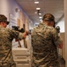 Two individuals wearing digital camouflage uniforms (consistent with USMC MARPAT pattern) are participating in what appears to be an augmented or mixed reality training exercise in an indoor corridor. Both are equipped with head-mounted display devices and are holding simulated weapon controllers with cables attached. The setting appears to be a training facility or office building repurposed for simulation-based tactical training.