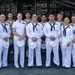 A formal group photograph of approximately 14 U.S. military personnel posing together in front of what appears to be a naval vessel or military facility. The group includes members in multiple uniform types: Navy dress whites, Navy working khakis/service uniforms, and Marine Corps service uniforms. Several sailors are holding white combination covers (hats) in their hands, suggesting a recent or upcoming ceremony.