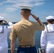 Three military personnel are standing on the deck of a naval vessel, saluting toward open water. The group consists of two U.S. Navy sailors in white dress uniforms flanking a U.S. Marine in tan service uniform. They are photographed from behind against a backdrop of blue water, distant shoreline, and partly cloudy sky.