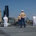 Military personnel are standing at parade rest on the flight deck of a naval vessel facing a city skyline, likely Seattle based on the distinctive skyscrapers visible in the background. The group includes both U.S. Navy sailors in white dress uniforms and at least one U.S. Marine in tan/khaki service uniform with blue trousers. This appears to be a formal 'manning the rails' ceremony, typically performed during port arrivals.