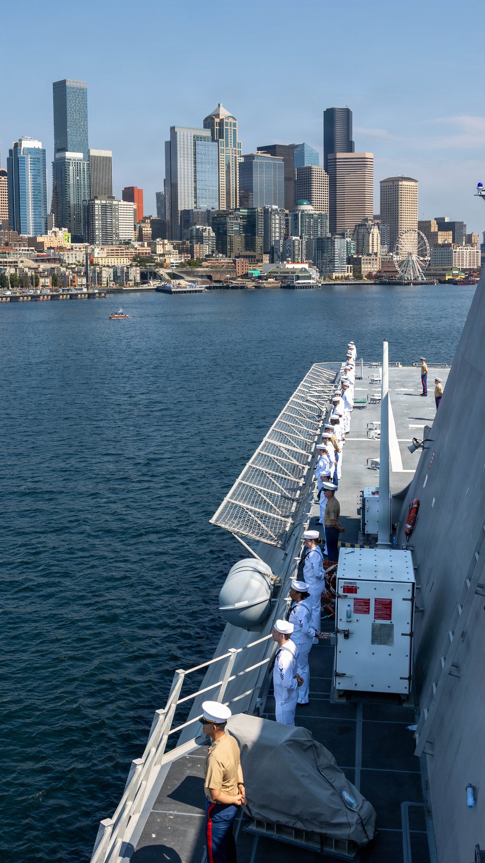 A U.S. Navy warship is manning the rails as it approaches or departs a port, with sailors and at least one Marine in dress uniforms standing at attention along the ship's deck. The city skyline visible in the background, including a prominent Ferris wheel and distinctive skyscrapers, is consistent with Seattle, Washington. The scene appears to be a formal naval arrival or departure ceremony, likely during Fleet Week or a similar public event.