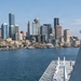 A U.S. Navy warship is manning the rails as it approaches or departs a port, with sailors and at least one Marine in dress uniforms standing at attention along the ship's deck. The city skyline visible in the background, including a prominent Ferris wheel and distinctive skyscrapers, is consistent with Seattle, Washington. The scene appears to be a formal naval arrival or departure ceremony, likely during Fleet Week or a similar public event.